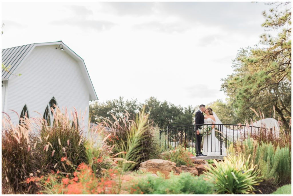Portrait of a bride and groom kissing on a bridge in front of a white wedding barn in Texas